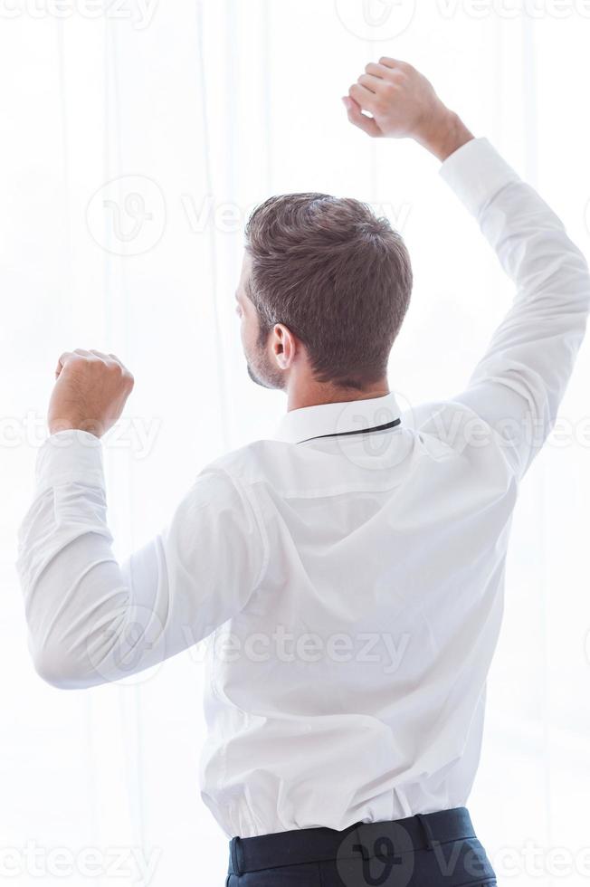 Time to relax. Rear view of man in shirt and tie standing against window and raising his arms photo