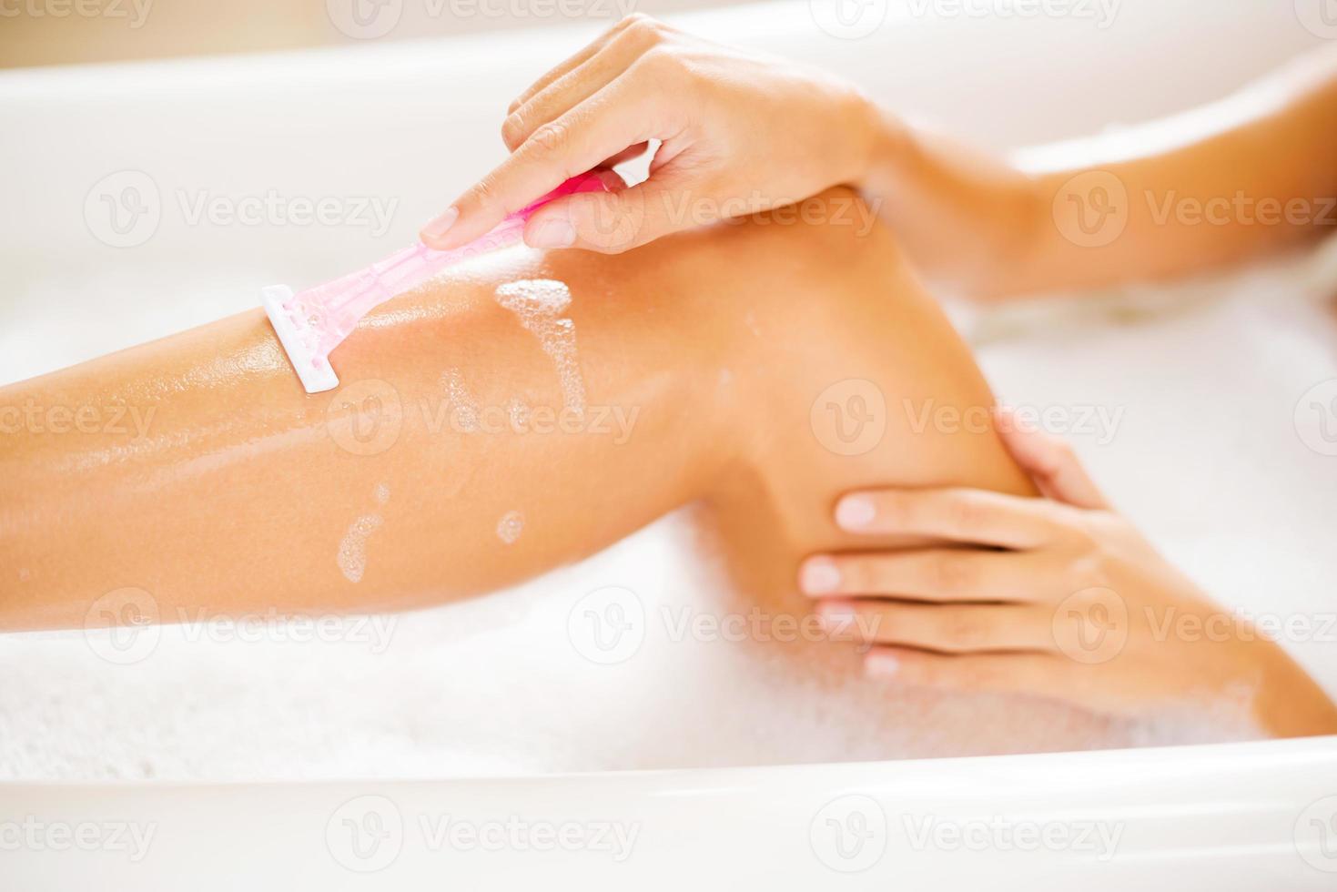 Making her legs smooth. Side view of young woman shaving her legs while lying in bathtub photo