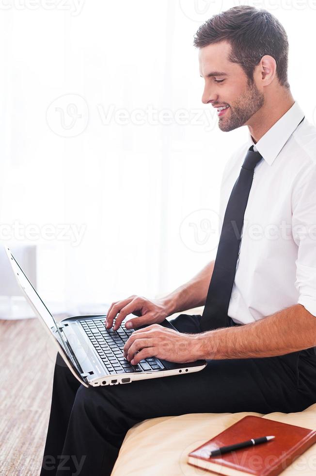 Staying connected. Side view of handsome young man in shirt and tie working on laptop and smiling while sitting on the bed photo