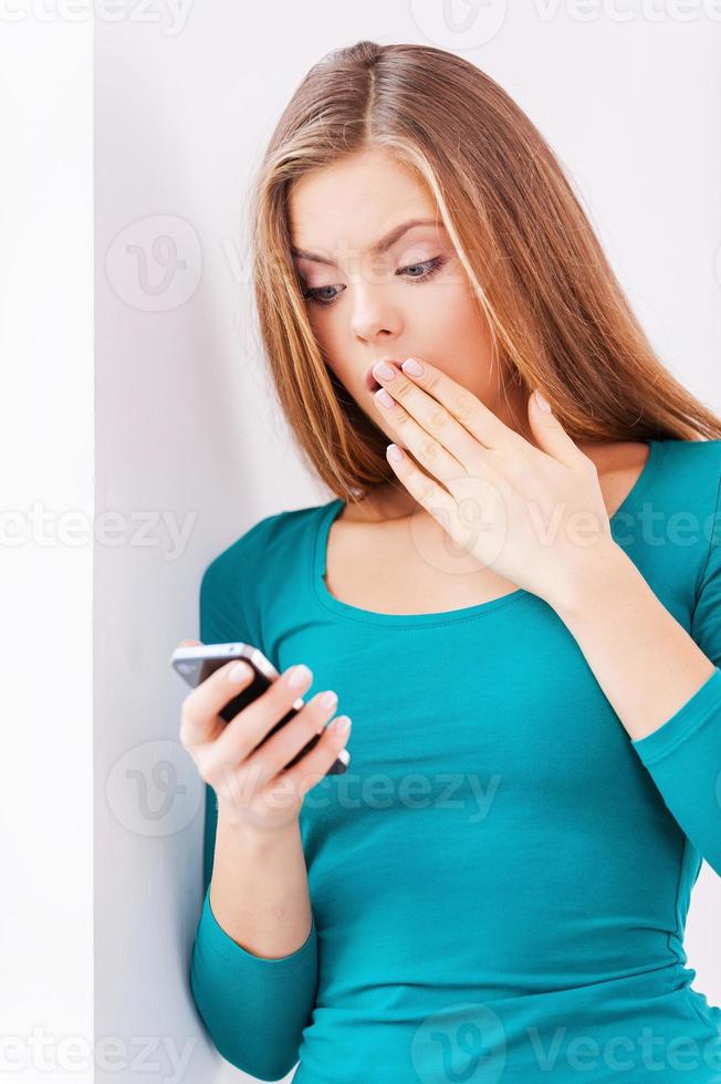 Shocking news. Surprised beautiful young woman holding phone in her hand while standing near window photo