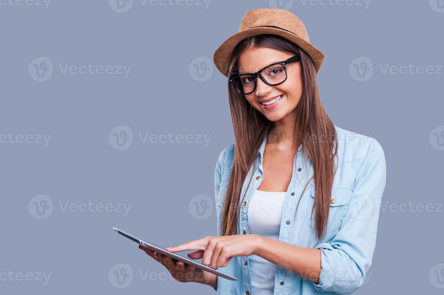 Examining new tablet. Beautiful young girl working on digital tablet and looking at camera while standing against grey background photo