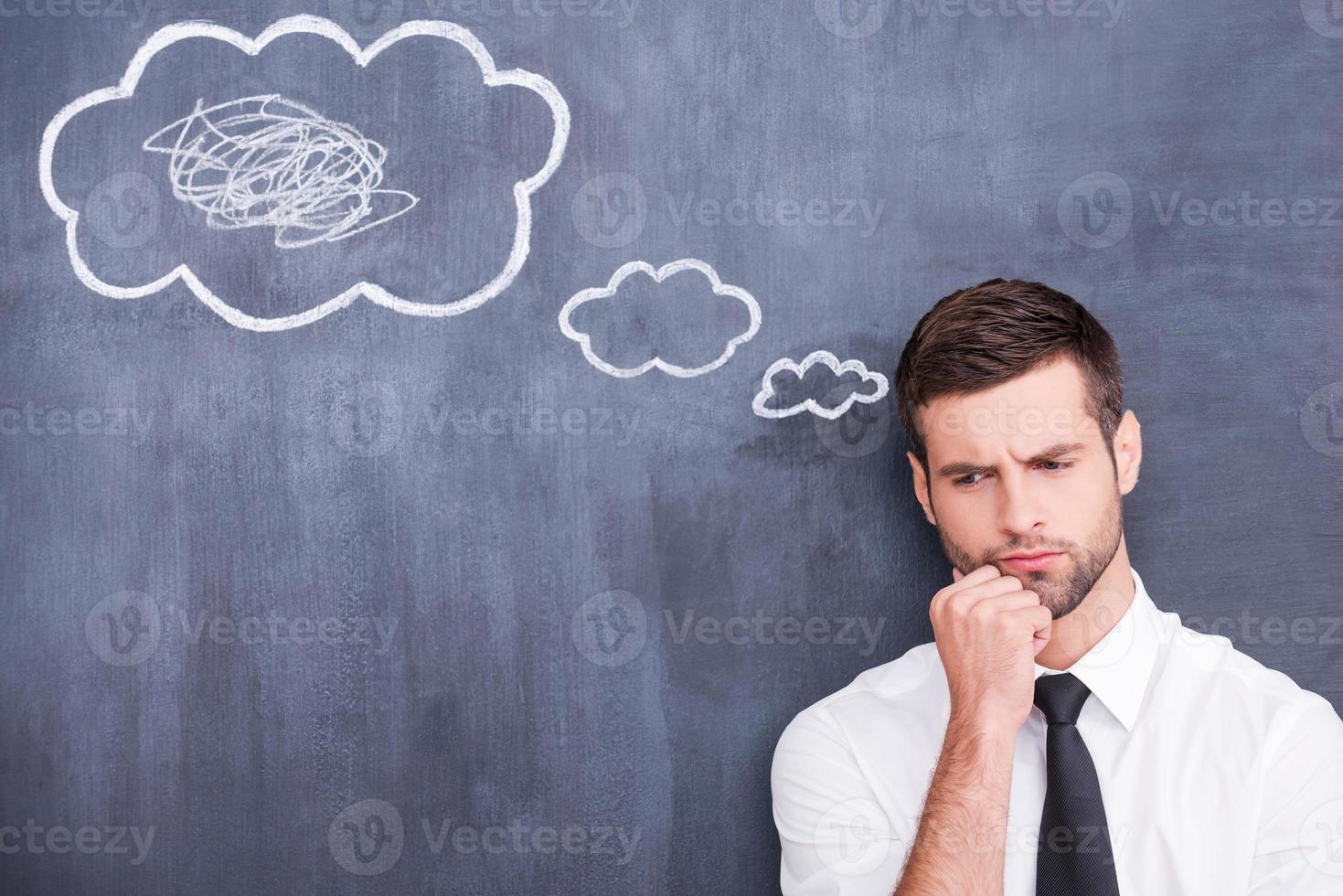 There is a mess in my head. Picture of confused handsome young man holding hand on chin and thinking while standing against cloud chalk drawing on blackboard photo