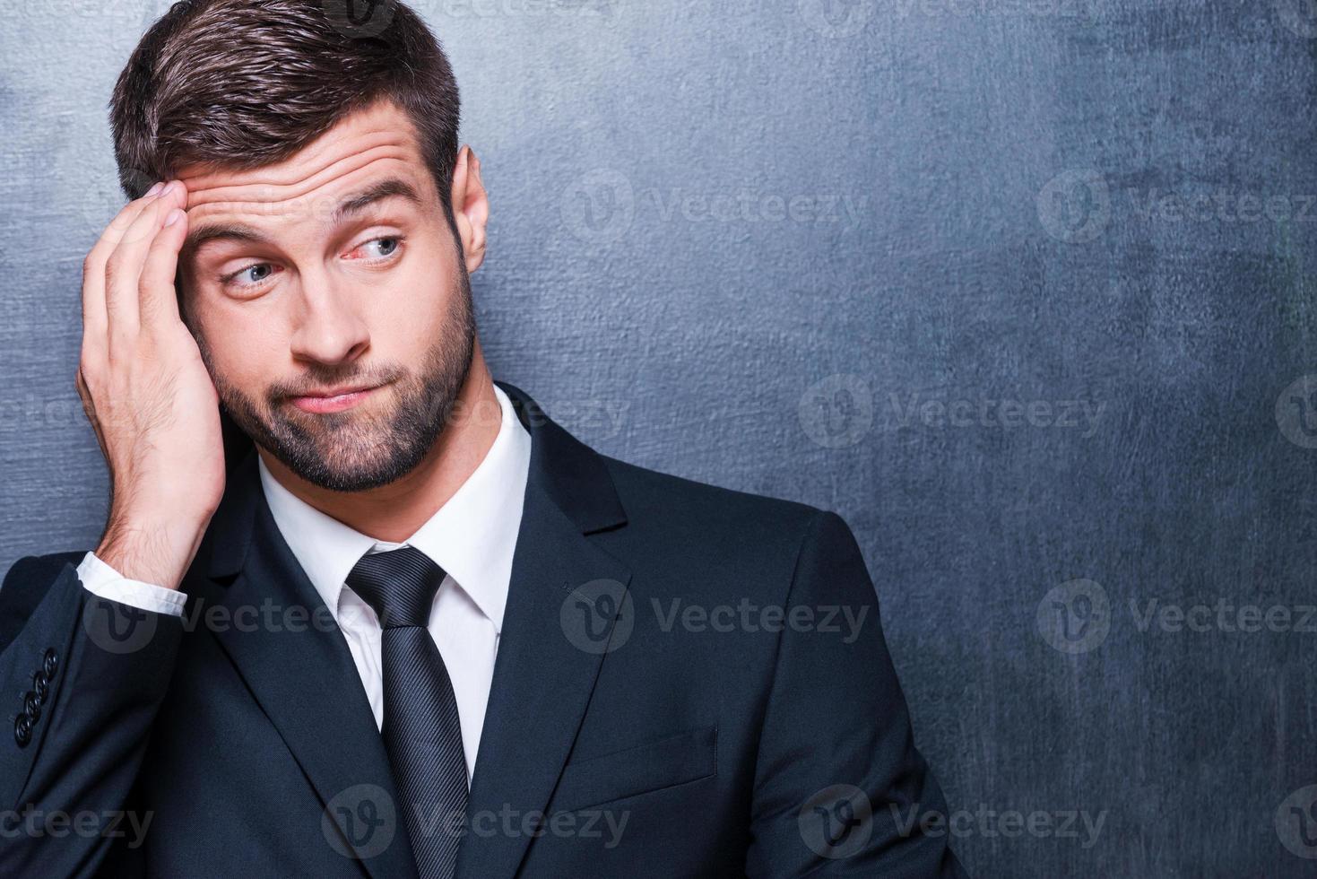 Feeling uncertain. Frustrated young man in formalwear holding hand on forehead and looking away while standing against blackboard photo