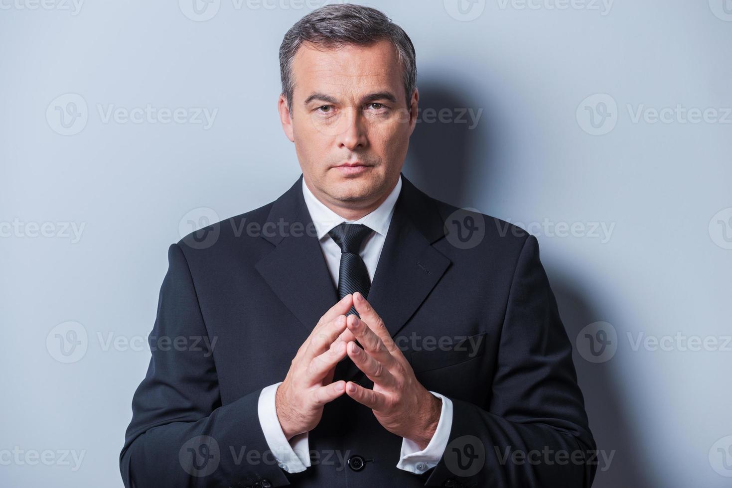 Confident business expert. Confident mature man in formalwear holding hands clasped and looking at camera while standing against grey background photo