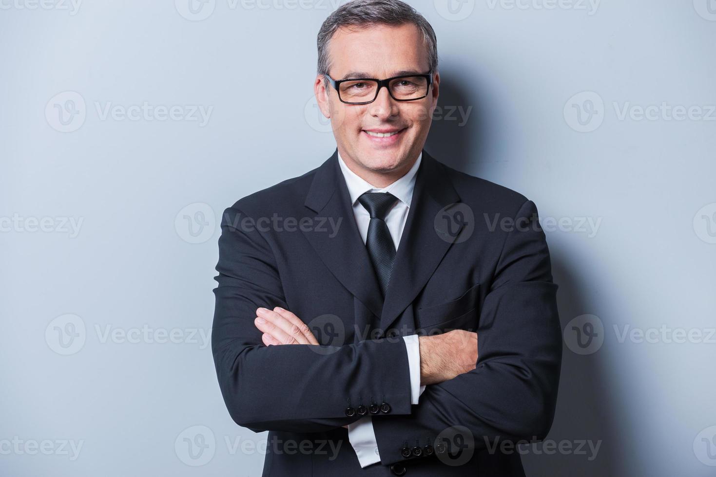 confiado y exitoso. retrato de un hombre maduro confiado en ropa formal y anteojos mirando a la cámara y sonriendo mientras se enfrenta a un fondo gris foto