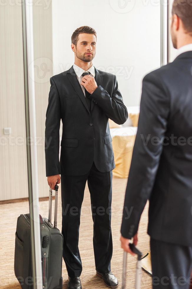 Ready for business trip. Confident young man in formalwear adjusting his necktie while standing against mirror in hotel room photo