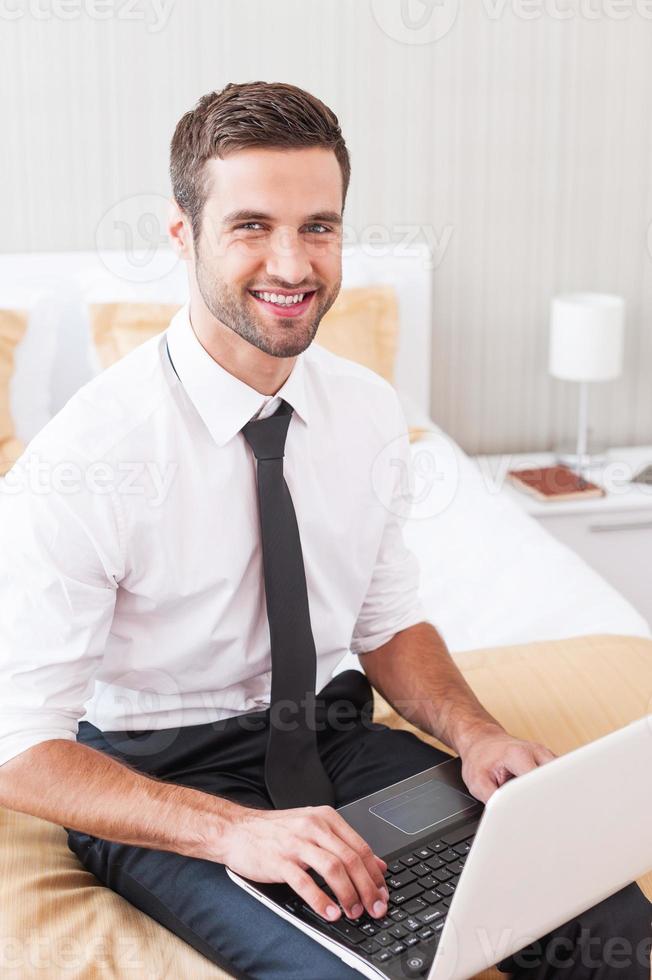 Staying connected anytime. Handsome young man in shirt and tie working on laptop and smiling while sitting on the bed in hotel room photo