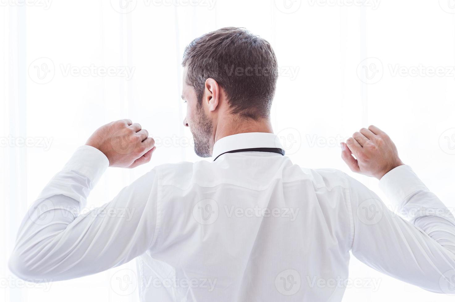 Time to relax. Rear view of man in shirt and tie standing against window and raising his arms photo
