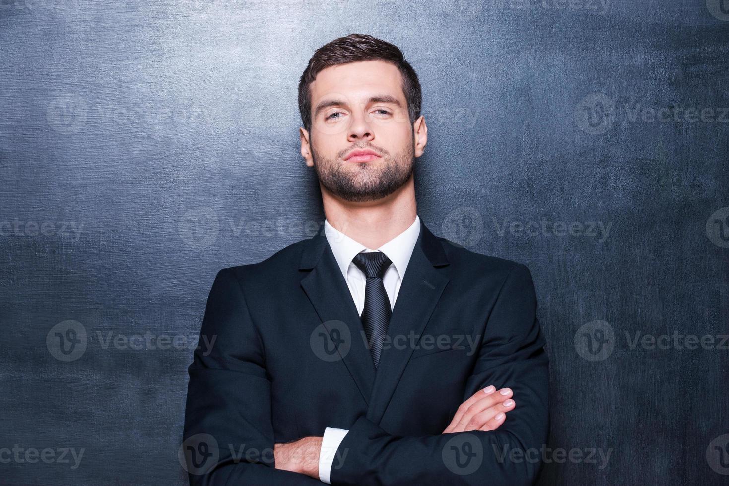 Confident business. Handsome young man in shirt and tie looking at camera and keeping arms crossed while standing against blackboard photo