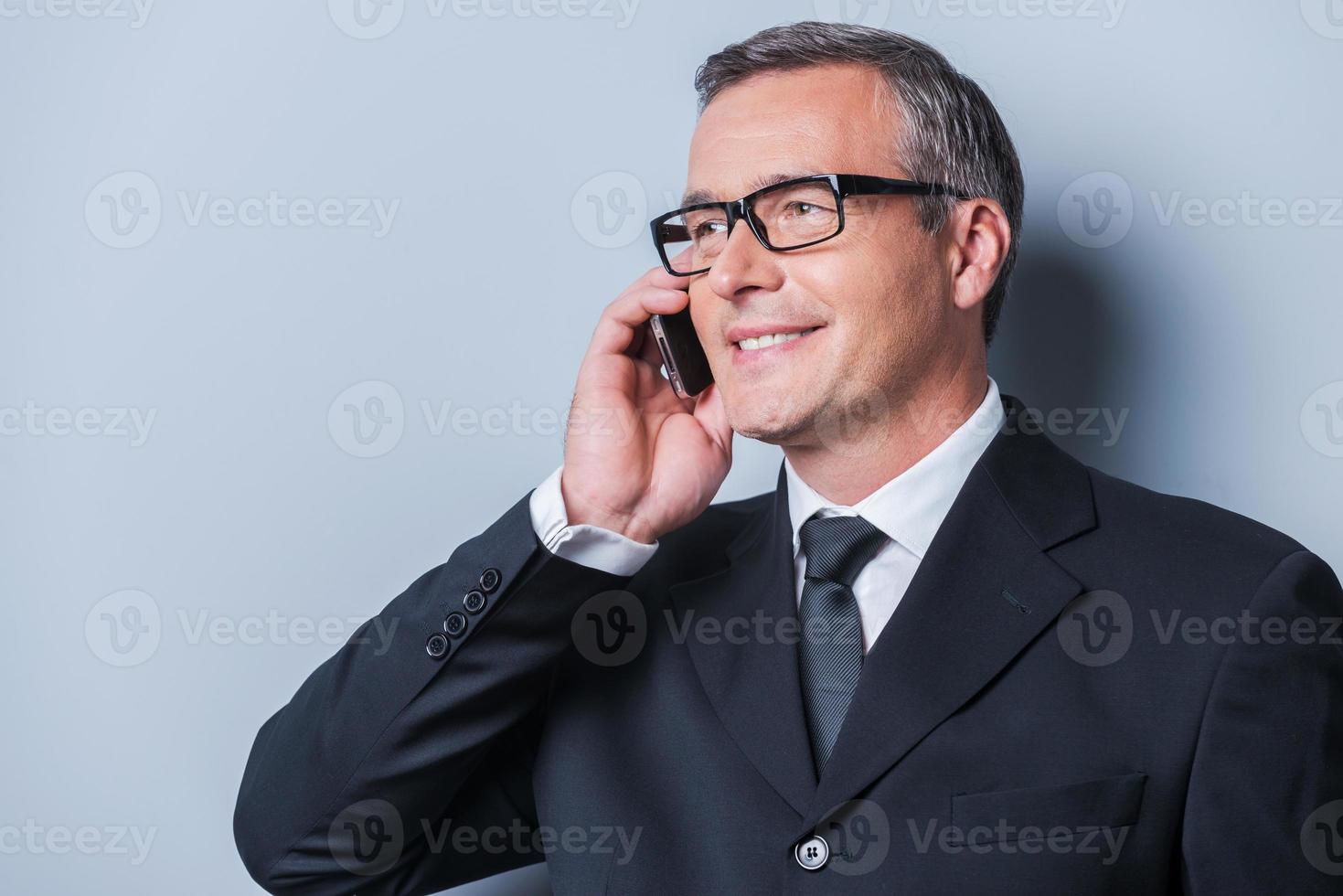 Good business talk. Portrait of confident mature man in formalwear talking on the mobile phone and smiling while standing against grey background photo