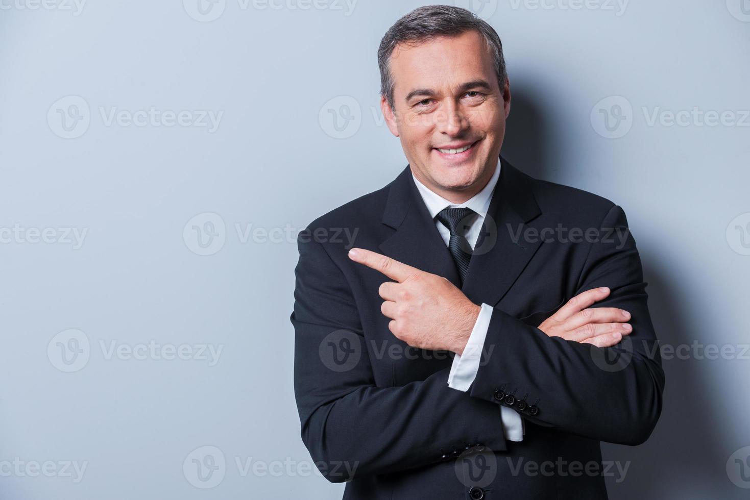 Advertising your product. Cheerful mature man in formalwear looking at camera and smiling while pointing away and standing against grey background photo