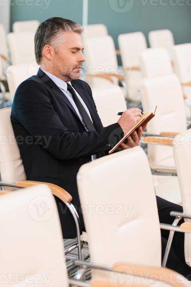 The one and only. Side view of concentrated mature man in formalwear writing something in note pad while sitting on the chair in empty conference hall photo