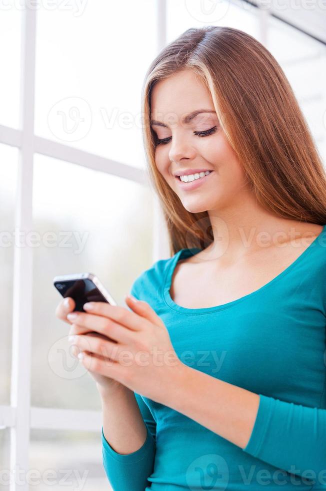 One message to warm your heart. Beautiful young woman holding phone in her hands while standing near window photo