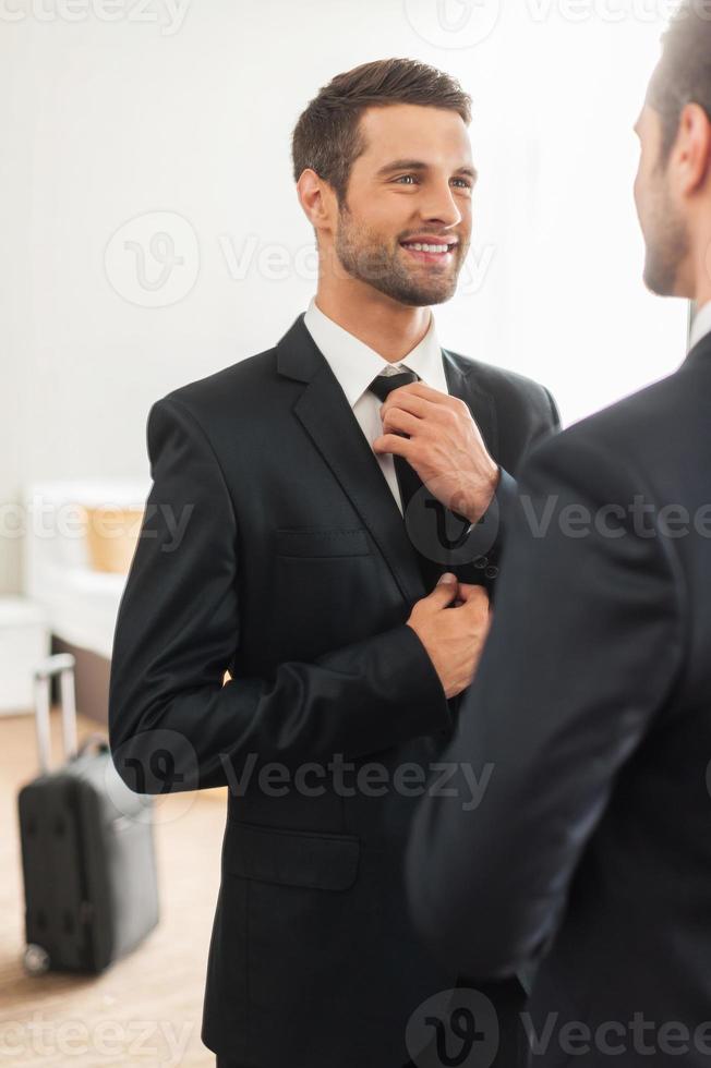Used to look perfect. Handsome young man in formalwear adjusting his necktie and smiling while standing against mirror in hotel room photo