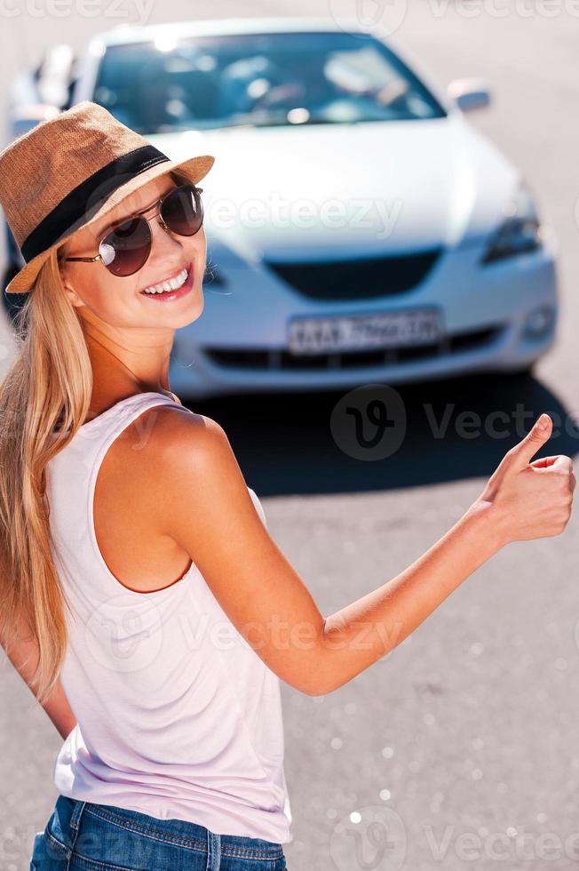 Hitching a ride. Beautiful young funky woman hitch-hiking on the side of the road and smiling with a car on background photo