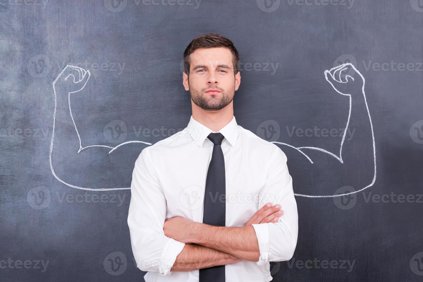 Hidden possibilities. Handsome young man in shirt and tie looking at camera and keeping arms crossed while standing against chalk drawing of muscular arms photo