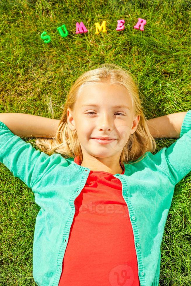 Enjoying summer time. Top view of cute little girl holding hands behind head and smiling while lying on the green grass with plastic colorful letters laying upon her head photo