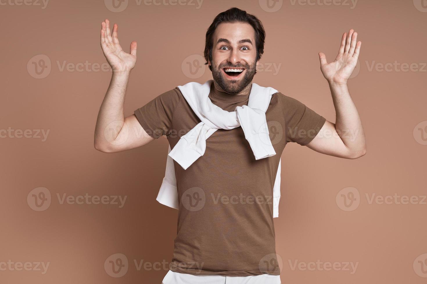 Excited young man gesturing and smiling while standing against brown background photo