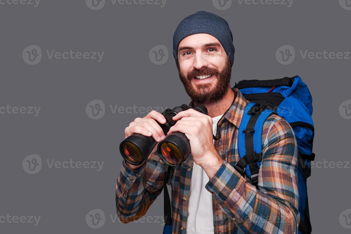 love hiking Handsome young bearded man with backpack holding binoculars and smiling while standing against grey background photo