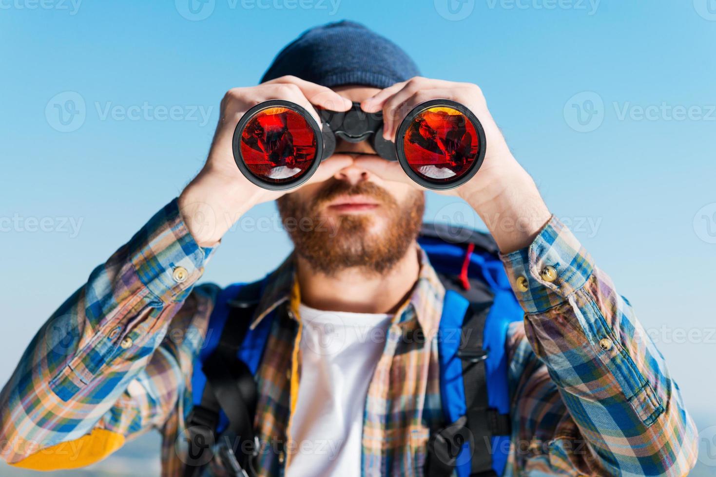 Opening of new horizons. Handsome young bearded man carrying backpack and looking through binoculars at camera photo