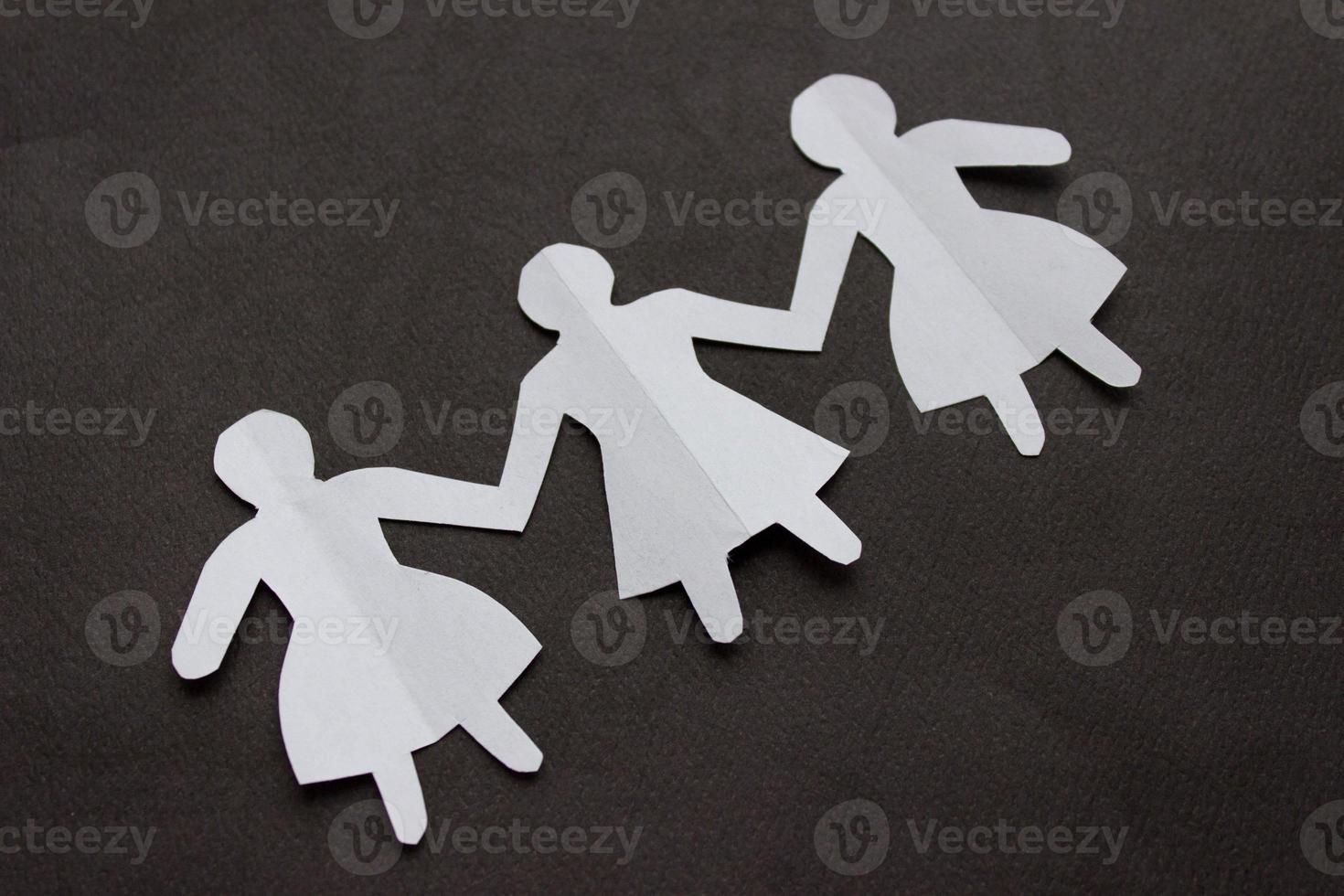 Figures of three girls in dresses holding hands, cut out of white paper. In the center of the photo on a black background