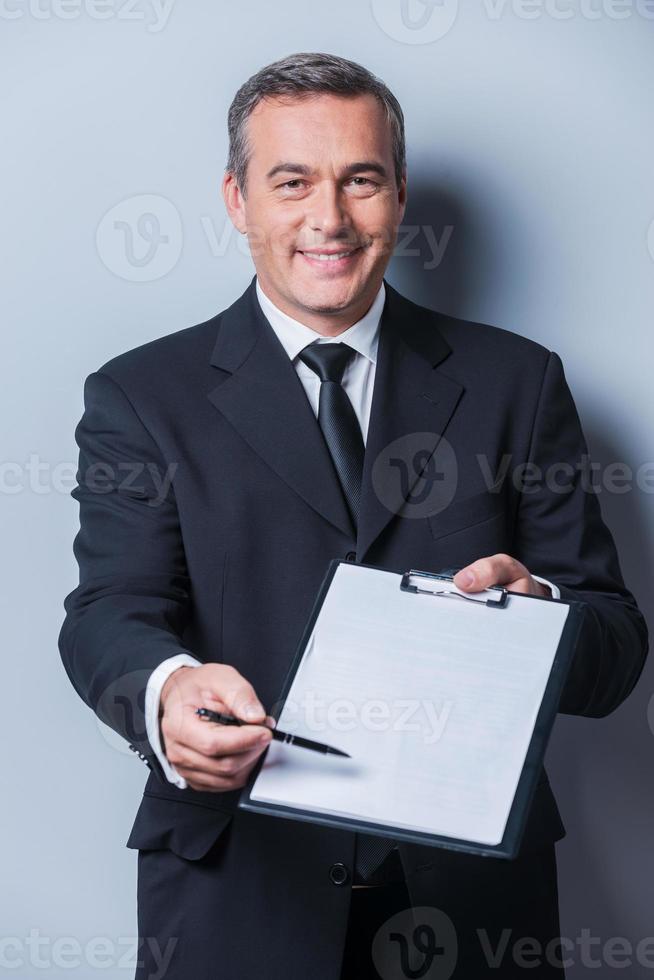 I need your signature here. Portrait of confident mature man in formalwear stretching out clipboard with paper and pointing it with pen while standing against grey background photo