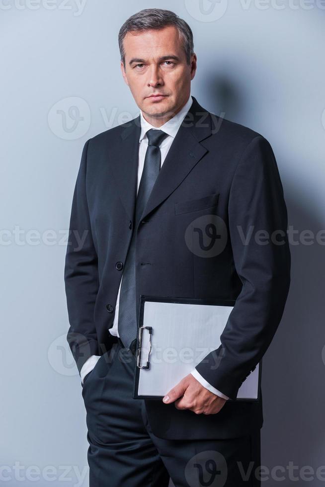 hombre de negocios confiado. hombre maduro confiado en ropa formal sosteniendo portapapeles y mirando a la cámara mientras está de pie contra el fondo gris foto