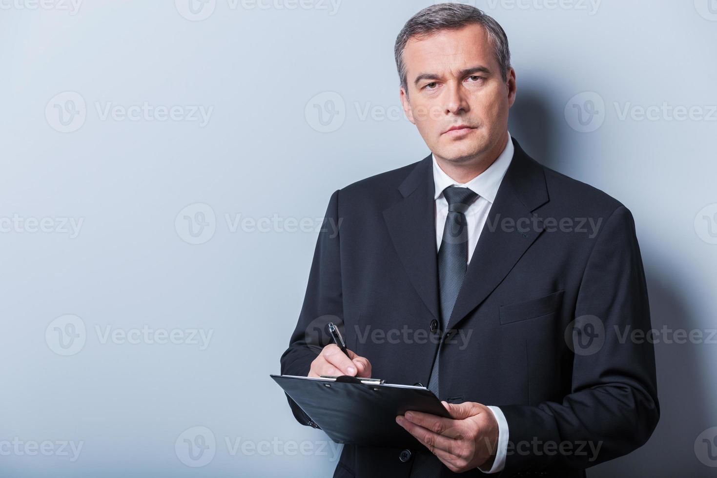 Making some urgent notes. Confident mature man in formalwear writing something in clipboard and looking at camera while standing against grey background photo