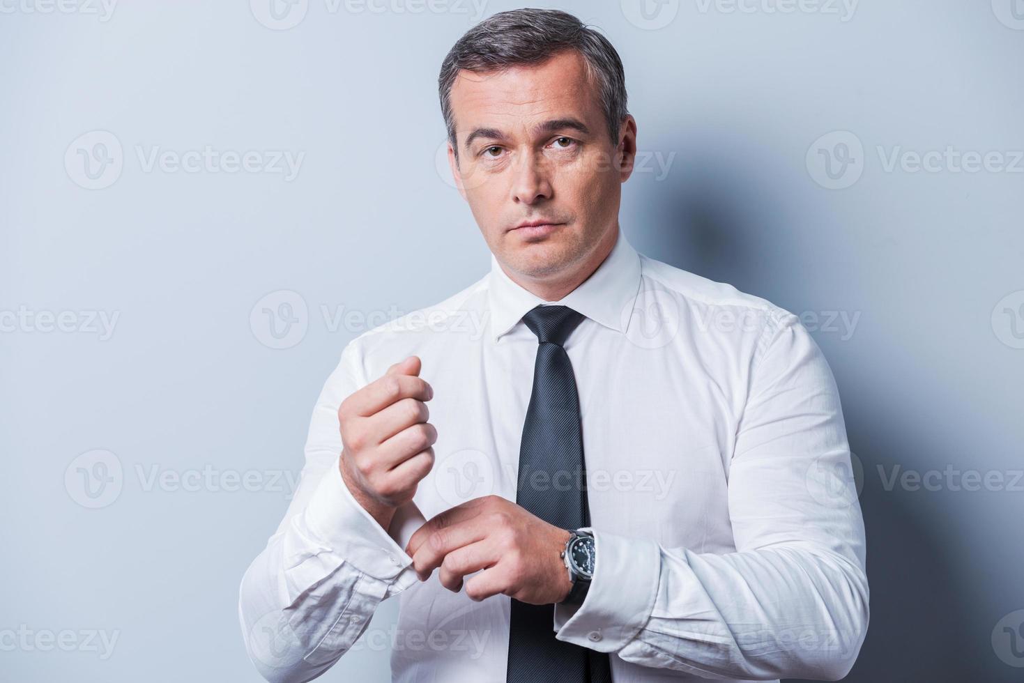 Making business look good. Confident mature man in shirt and tie adjusting his sleeves and looking at camera  while standing against grey background photo