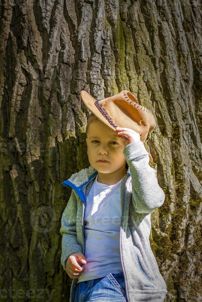 Cute boy posing in a cowboy hat in the woods by a tree. The sun's rays envelop the space. Interaction history for the book. Space for copying photo