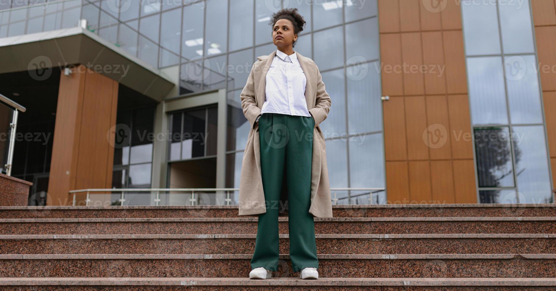 portrait of an African-American student in thought standing on the steps against the backdrop of an administrative city building, successful business deal concept photo