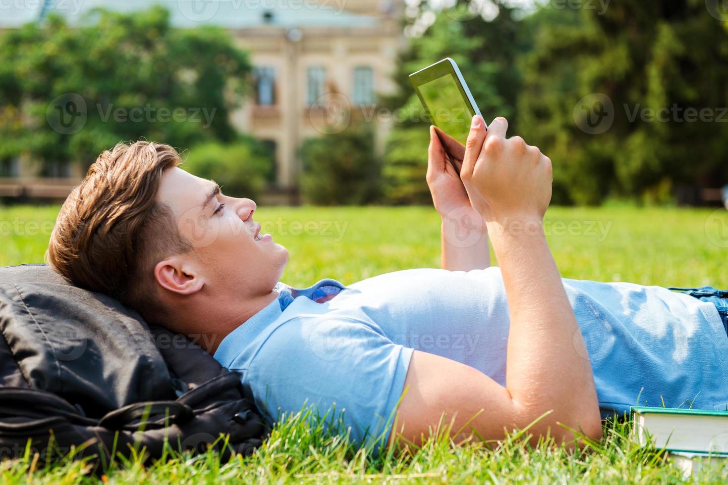 Finding a peaceful place to relax. Side view of handsome young man working on digital tablet while lying on grass photo