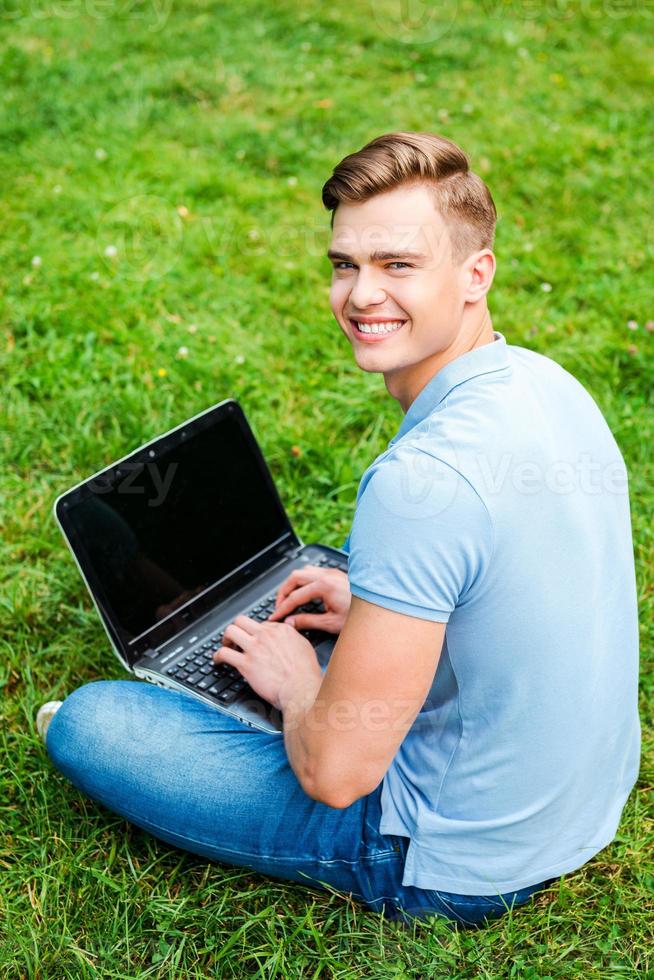Surfing the net wherever I want. Top view of happy young man looking over shoulder and smiling while sitting on the grass with laptop photo