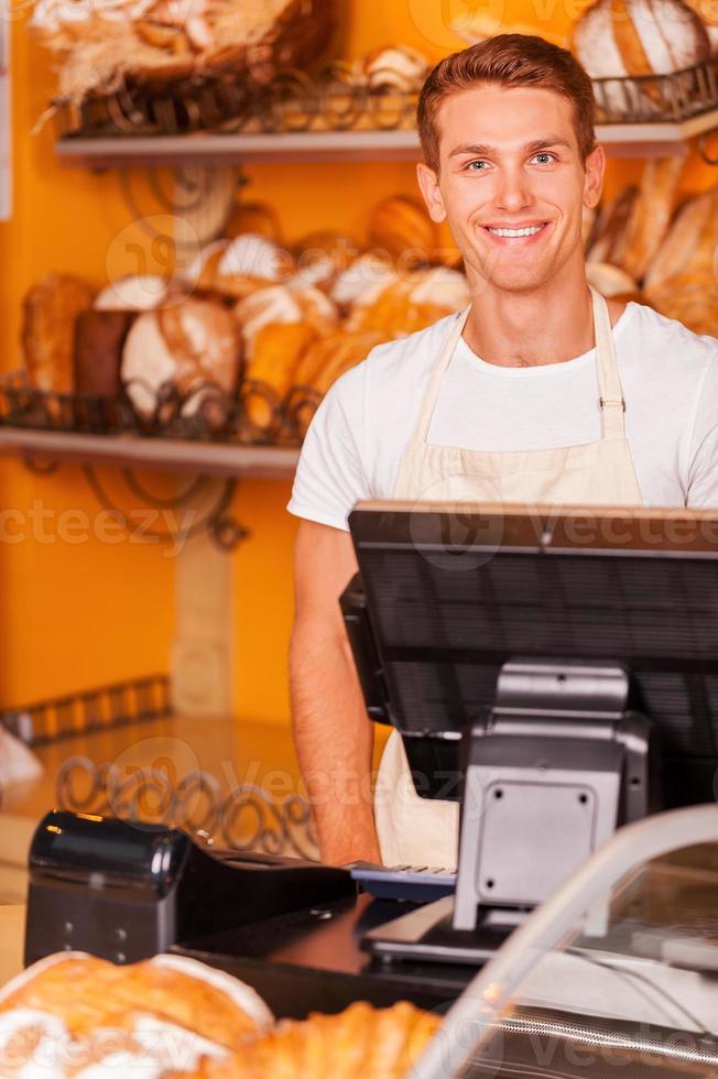 Confident cashier. Handsome young male cashier in apron standing near cash register and smiling while standing in bakery shop photo