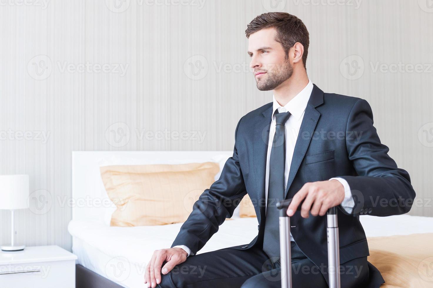 Ready to new business trip. Confident young businessman in formalwear carrying suitcase and smiling while sitting on the bed in hotel room photo