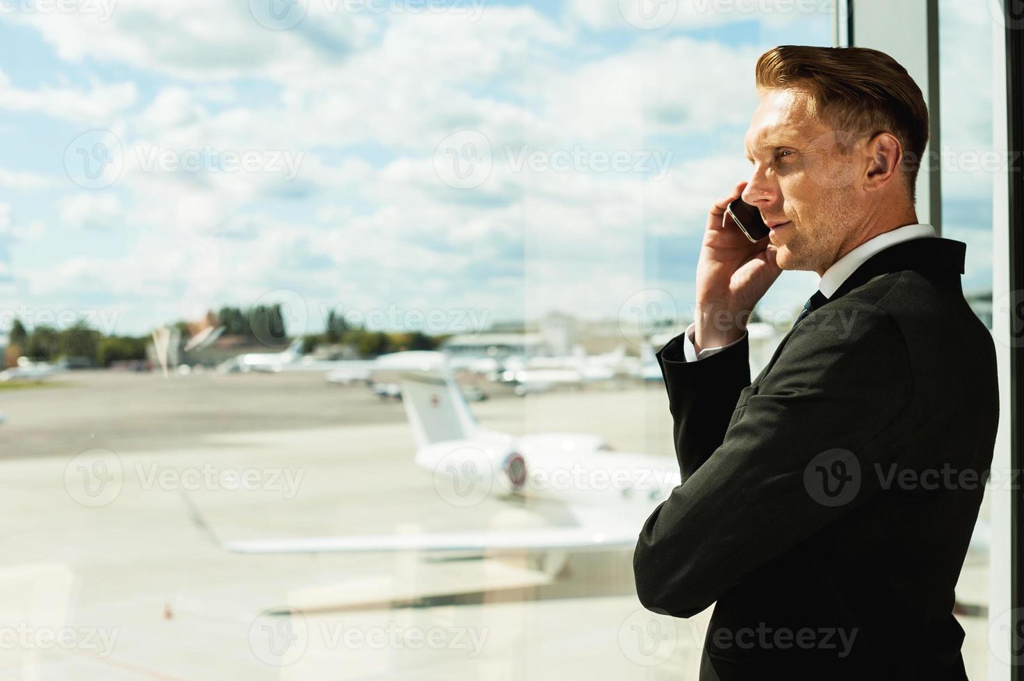 Businessman waiting for flight. Rear view of thoughtful businessman in formalwear looking through a window and talking on the mobile phone while standing in airport photo