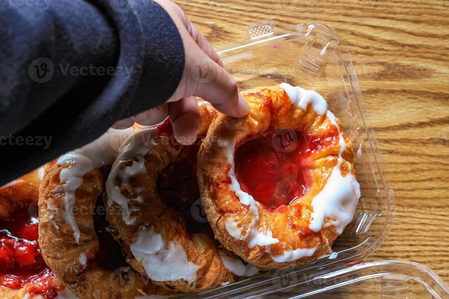 A man's hand is holding a Cherry filled Danish or Danish bread in a plastic box and placed it on a brown wooden table. photo