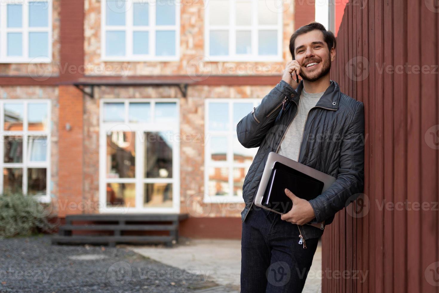 adult male businessman talking on a mobile phone leaning against a wooden fence next to a brick building photo