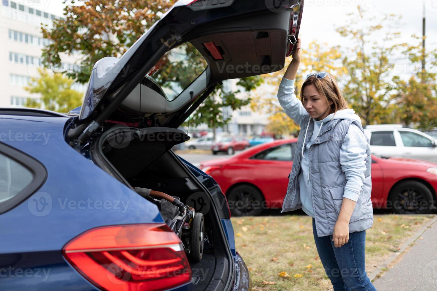 a young woman mother loaded a baby stroller into the car and stands near the trunk of the car photo