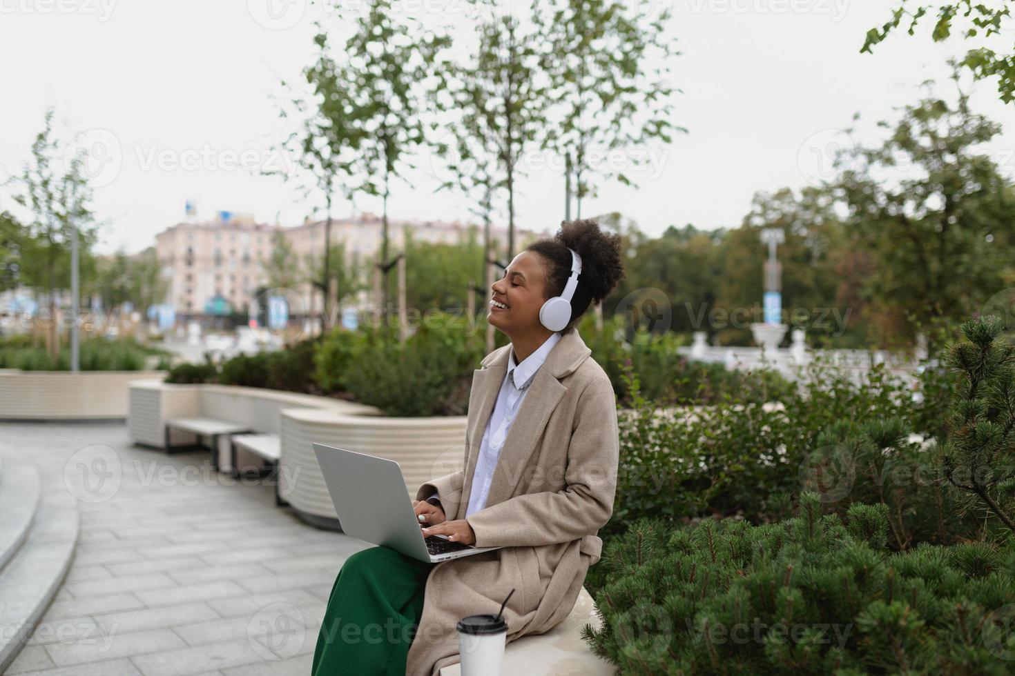 a young black student with a laptop sitting on a bench listening to music in headphones with pleasure closing her eyes photo