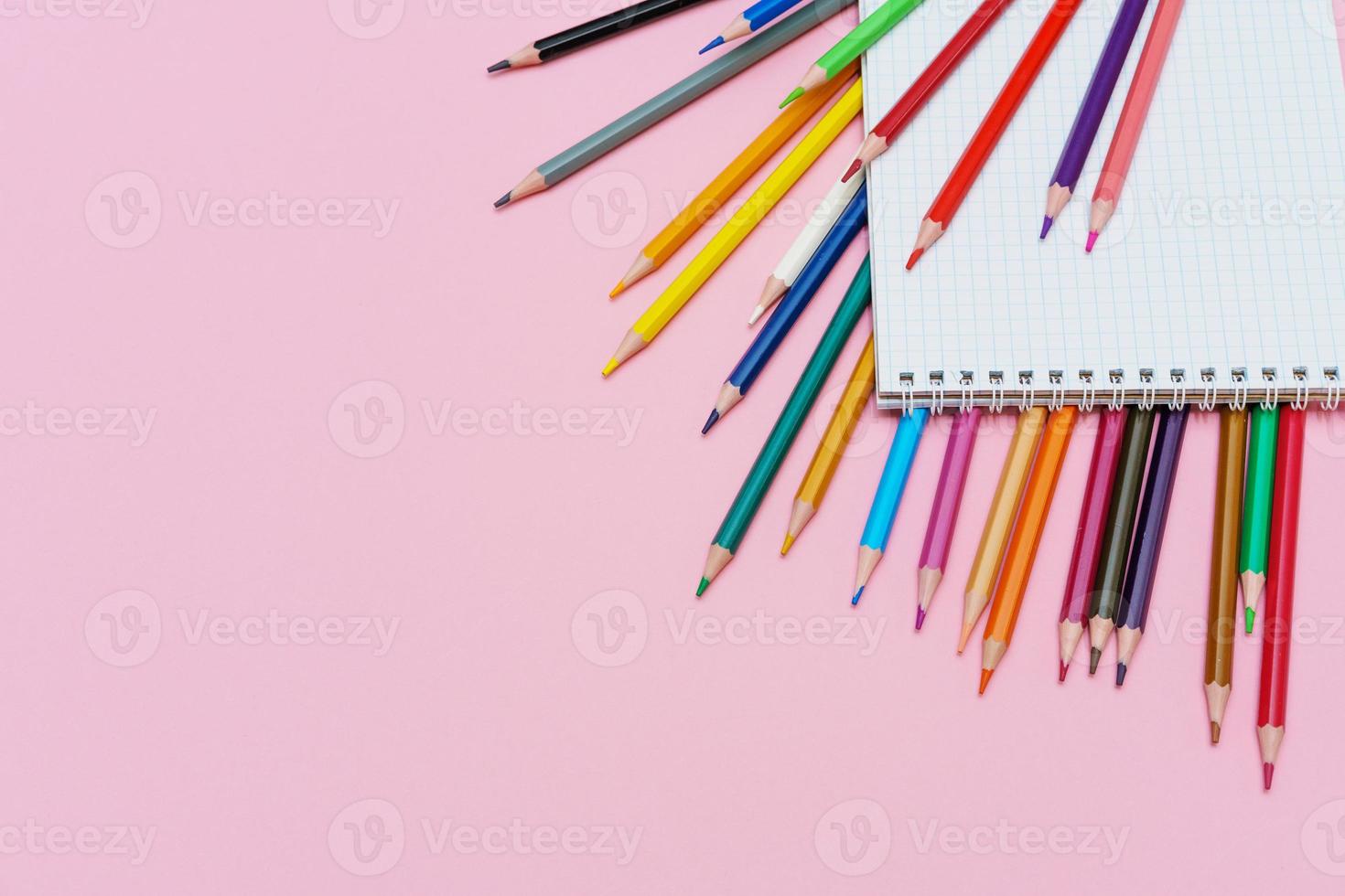Colored pencils for drawing and a notebook in a cage on a pink background. Back photo