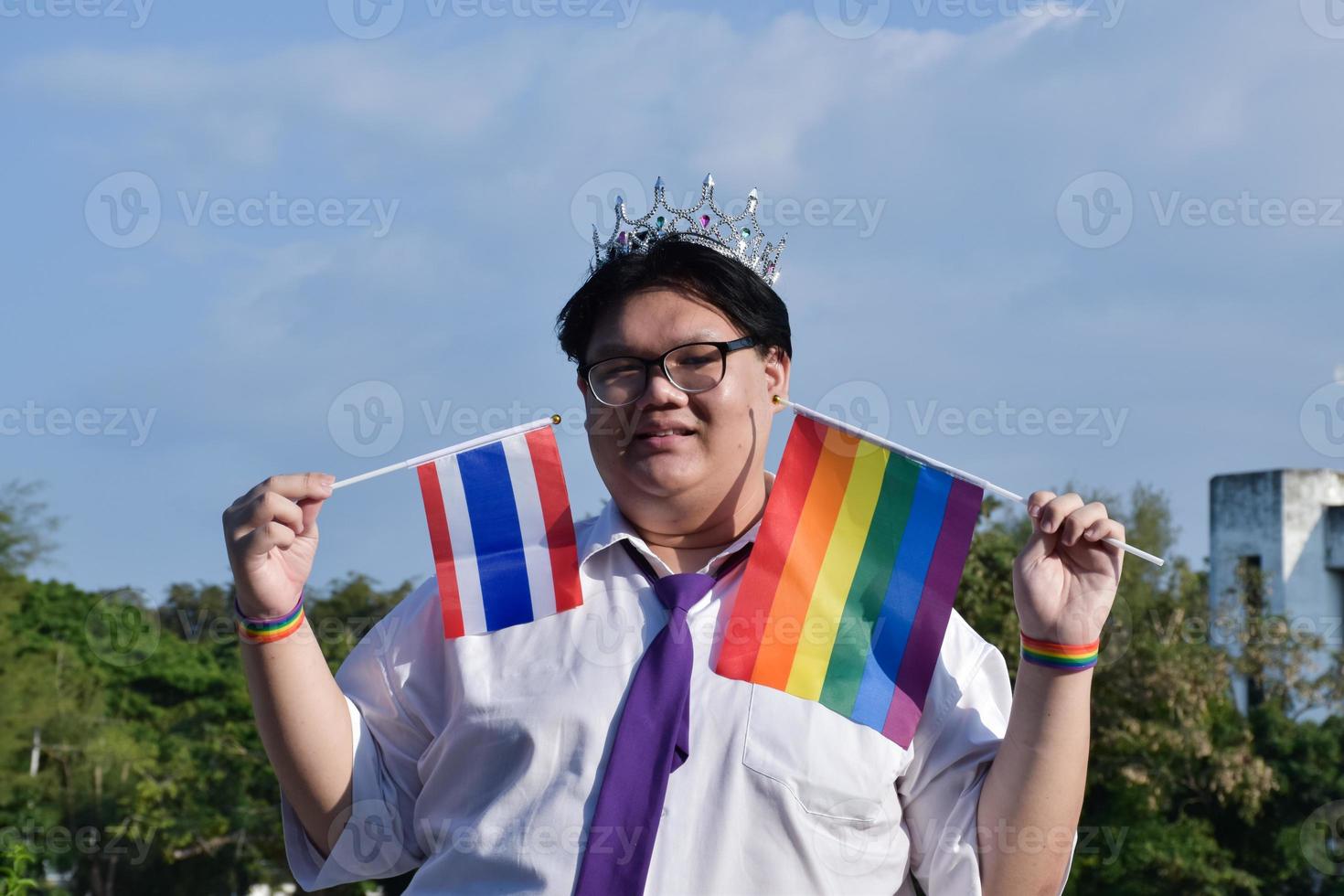 Thai boy holds rainbow flag and Thailand national flag and wears crown and violet neck tie and standing outside the building, soft and selective focus, concept for lgbtq celebration in pride month. photo