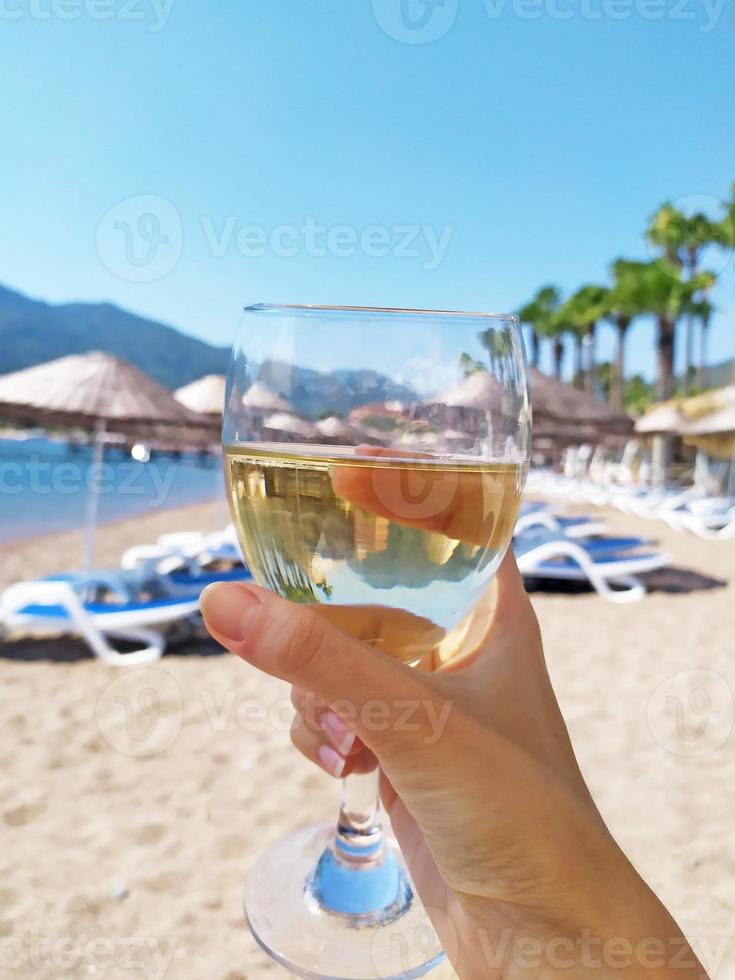 a glass of white wine in hand on the background of a sunny beach with sun loungers, umbrellas and palm trees photo