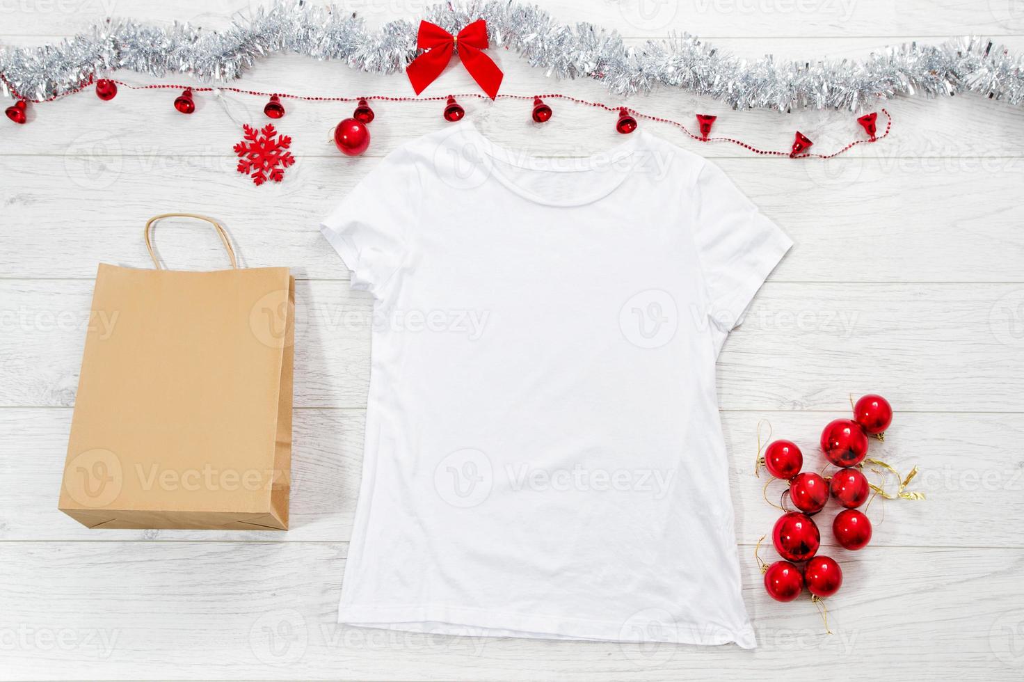 Close up white blank template t shirt with copy space paper bag. Christmas Holiday concept. Top view mockup t-shirt, star, white wooden background. Happy New Year decorations accessories. Xmas outfit photo