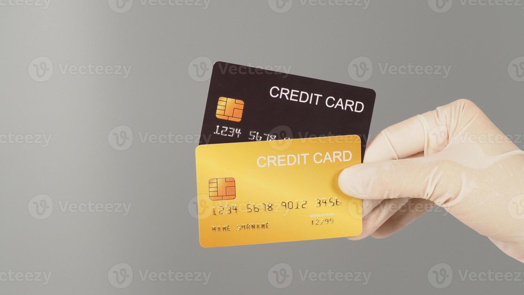 las manos sostienen dos tarjetas de crédito negras y doradas sobre fondo gris. la mano lleva un guante médico blanco. foto