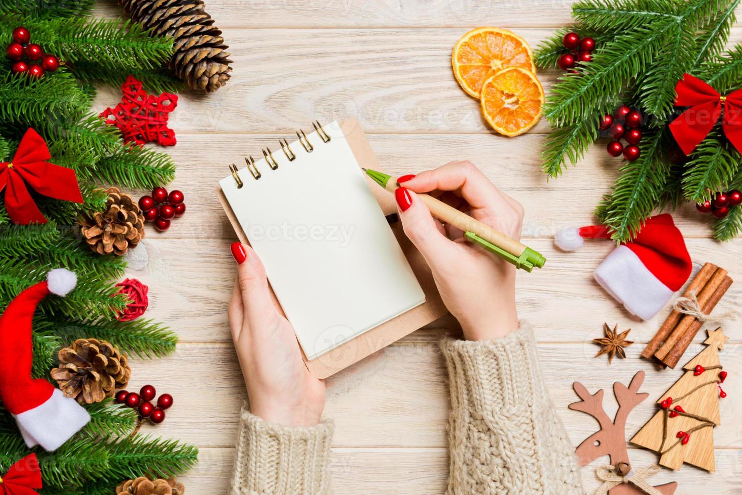 Top view of female hand writing in a notebook on wooden Christmas background. Fir tree and festive decorations. Wish list. New Year concept photo