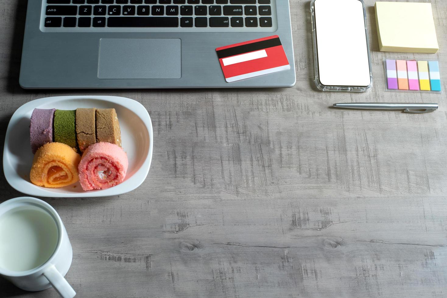 Top view of laptop, smartphone, credit card, cup of milk, jam roll, paper note, pen, on wooden table with business, commerce, finance, education concept and design photo