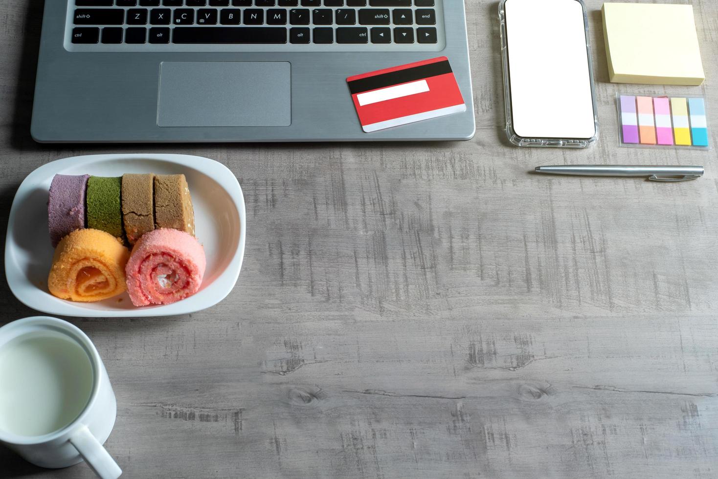 vista superior de la computadora portátil, teléfono inteligente, tarjeta de crédito, taza de leche, rollo de mermelada, nota de papel, pluma, sobre una mesa de madera con negocios, comercio, finanzas, concepto de educación y diseño foto