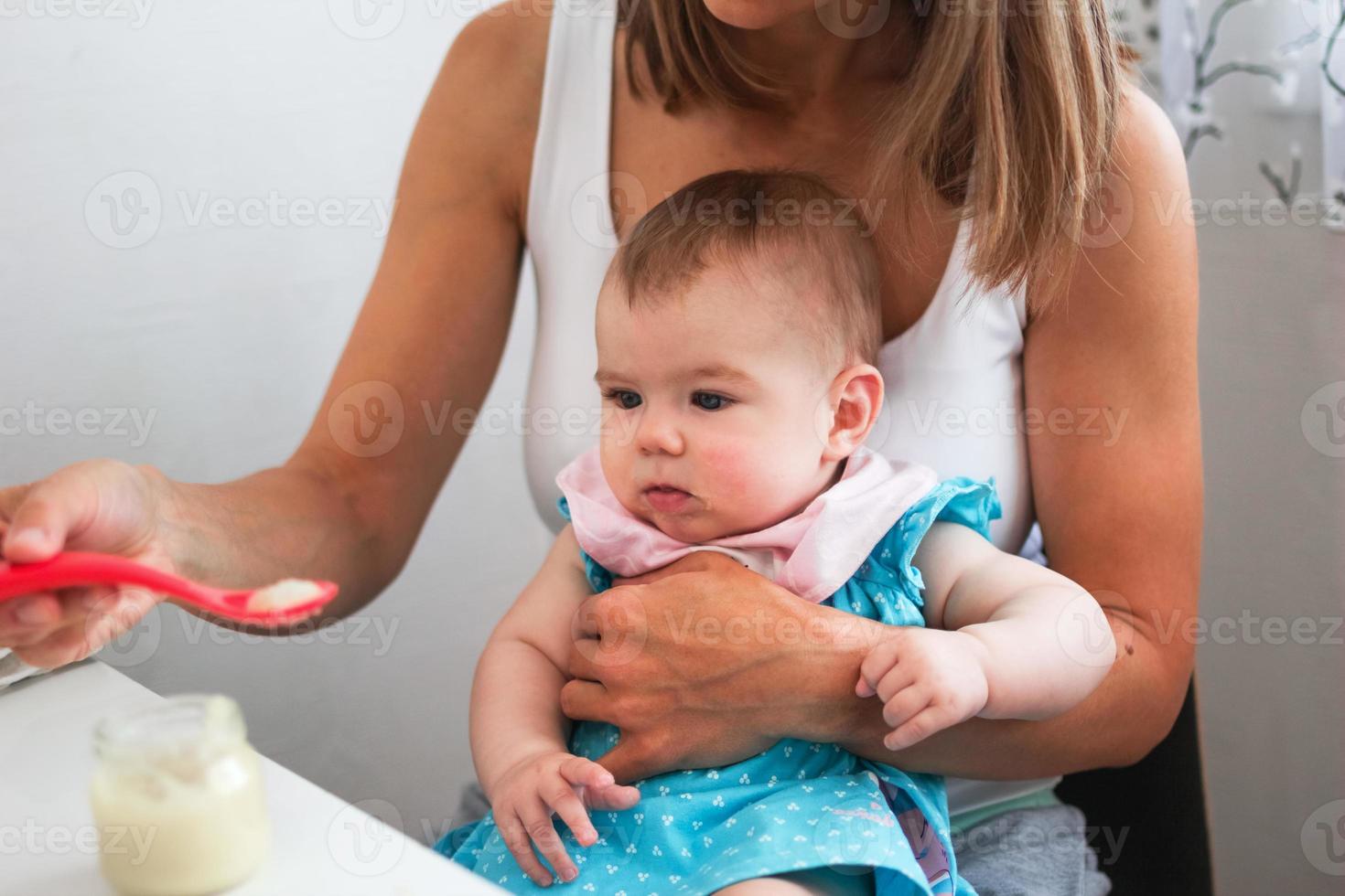 Mother feeding cute baby girl vegetable puree from a spoon on kitchen. Healthy eating nutrition for little kids photo