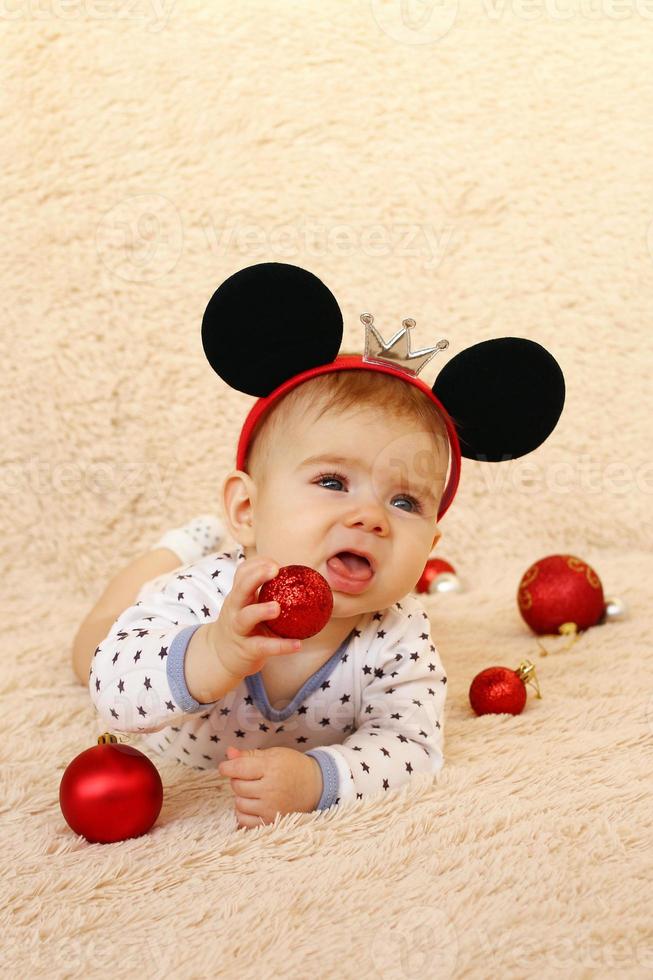 adorable niñita con orejas de ratón está acostada sobre una manta beige y jugando con bolas de Navidad rojas y brillantes. foto